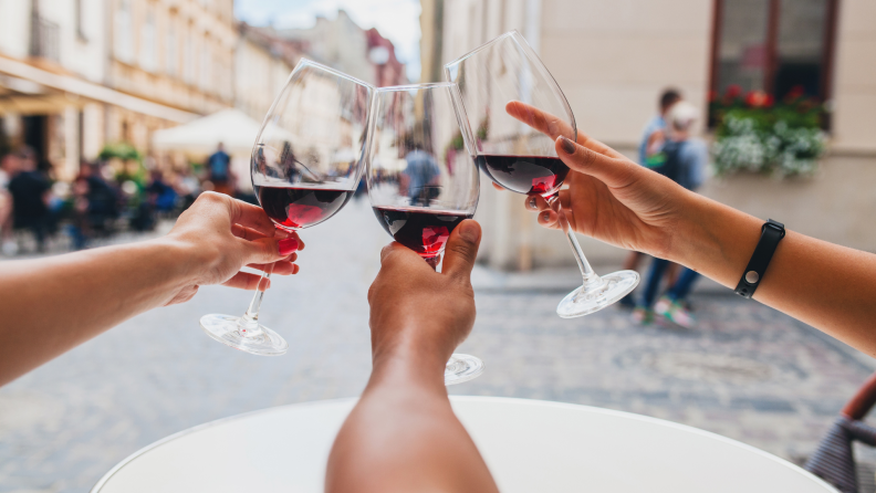 Three people toast with glasses of red wine at an outdoor café, with a lively European street in the background.