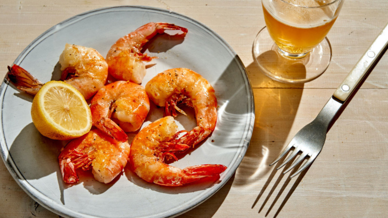 In a well-lit room, a plate of cooked shrimp is on display, accompanied by a fork and a glass of beer.