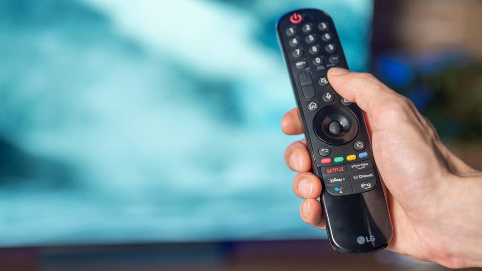 Hand holding a TV remote in front of a blurry TV.