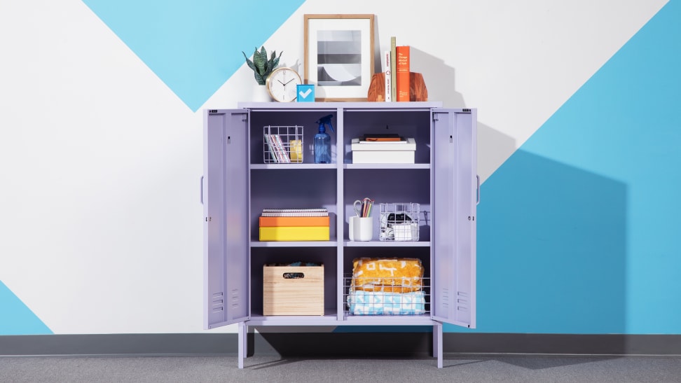 A lilac Mustard Made locker against a wall with its doors open and shelves full of housewares. The top is set with books, photos, and a plant.