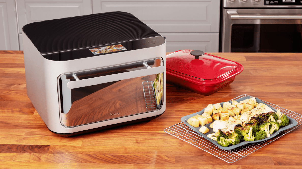 The Brava Glass Oven on a wooden countertop next to a plate of roasted vegetables and a red cassarole dish.