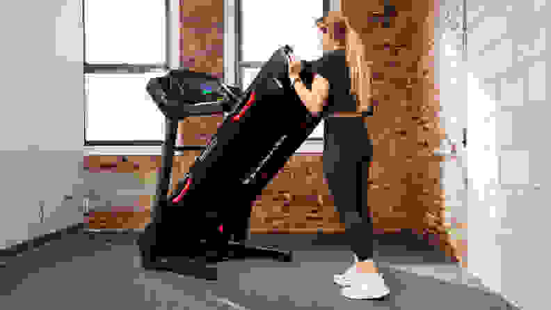 A woman lifts up the underside of a Bowflex BXT8J as if to fold it.