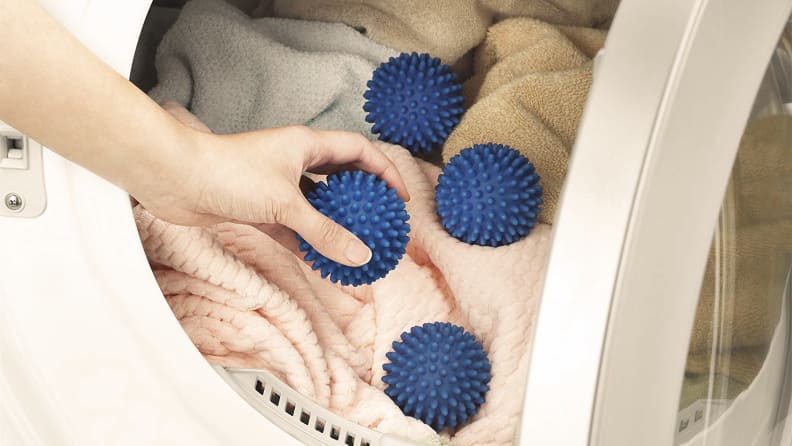A hand model inserts four blue Whitmor dryer balls into a dryer, along with a load of blankets and soft-looking towels.