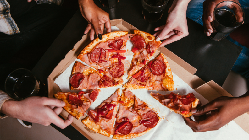 People reaching into pizza box to extract slices of pizza topped with assorted meats.