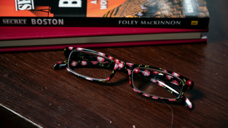 Pair of single pink, white and black rectangular reading glasses on wood table next to a stack on books.