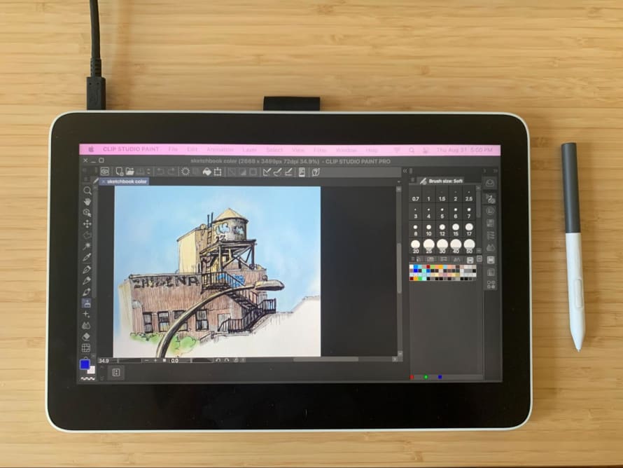 The image shows the Wacom One 13 Touch drawing tablet with Clip Studio Paint pulled up on the screen, showing a digital painting—next to it sits Wacom’s digital pen.