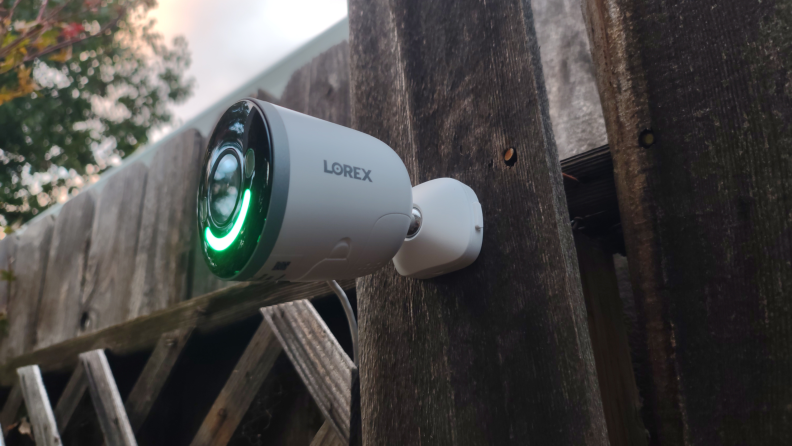 Lorex 4K Spotlight mounted on wooden fence outdoors with green LED light.
