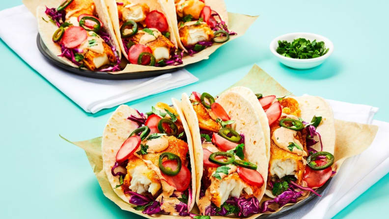 Three fish tacos arranged on a square white plate, accompanied by a second plate of tacos in the background.