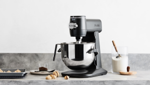 A dark grey GE Smart Stand Mixer sits on a grey counter surrounded by baked goods and glass canister of sugar with a measuring cup in it.