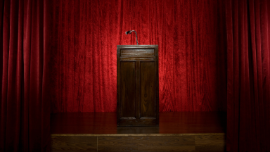 An image of a wooden podium sits on a stage, surrounded by red velvet curtains.