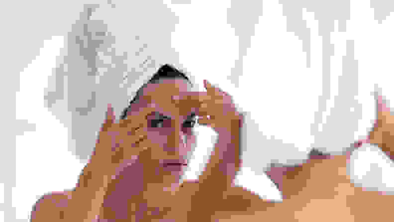 A person wearing a towel on their head, looking into a mirror, and touching their forehead.