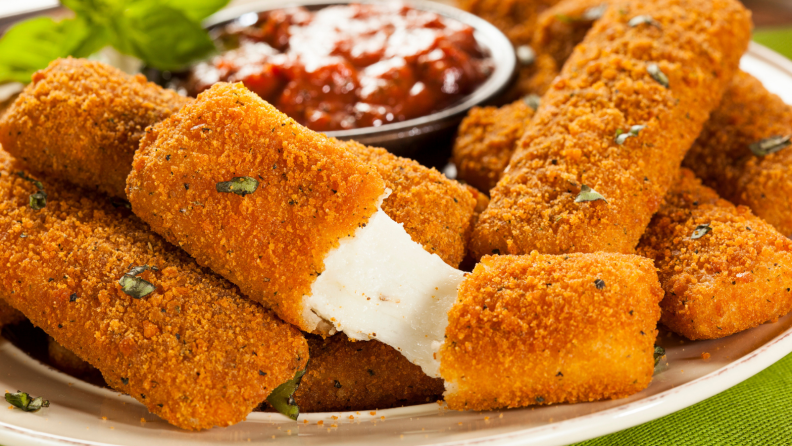 A plate of fried mozzarella sticks with a side of marinara sauce; one mozzarella stick is being pulled in the center to reveal the gooey filling.