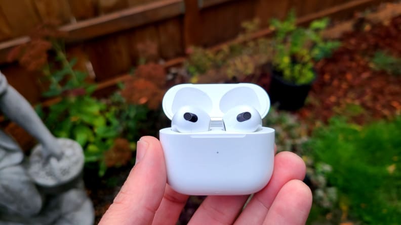 The all-white AirPods (3rd generation) sit in an open case before a fall background that includes a stone statue, leaves, green grass, and wood chips.