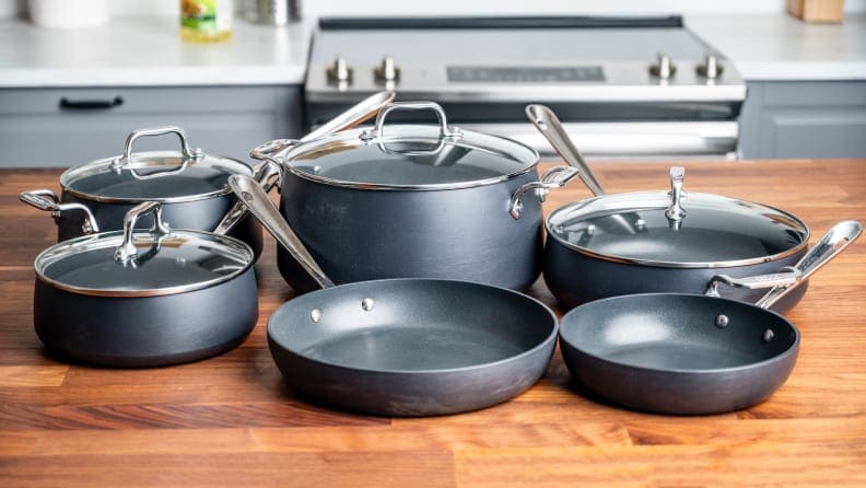 A collection of steel grey All-Clad pots and pans on a kitchen island in the foreground, a stovetop and counter in the background.