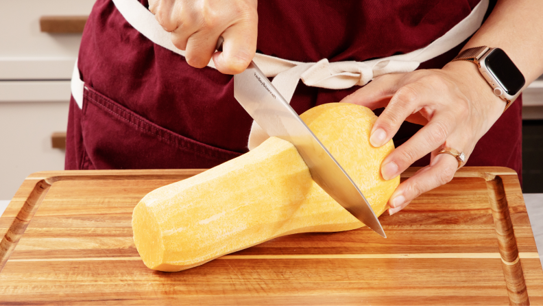 Person using knife to cut butternut squash in half horizontally on top of wooden cutting board.