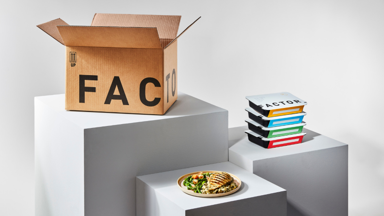 Against a white backdrop, there's a box of Factor pre-made meal delivery service on the left. In the middle, there's a plate of chicken dish and to the right, there are four Factor meals stacked on top of each other.