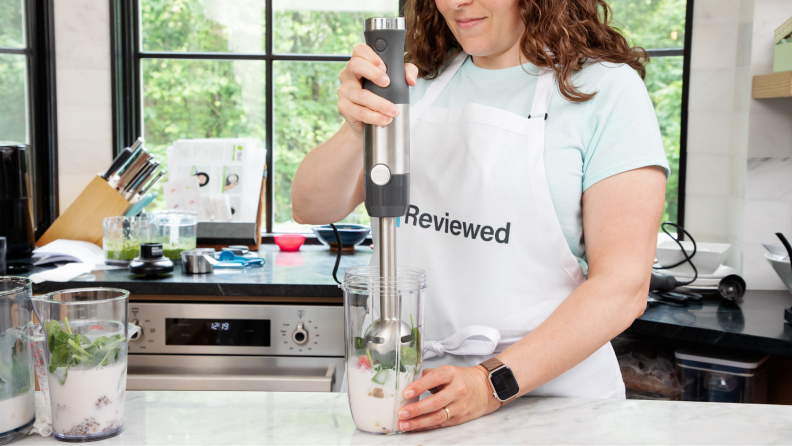Person with apron on stands at kitchen countertop while using GE Immersion Blender to blend smoothie ingredients inside of plastic cup.
