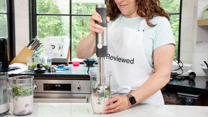 人与围裙站在厨房countertop while using GE Immersion Blender to blend smoothie ingredients inside of plastic cup.