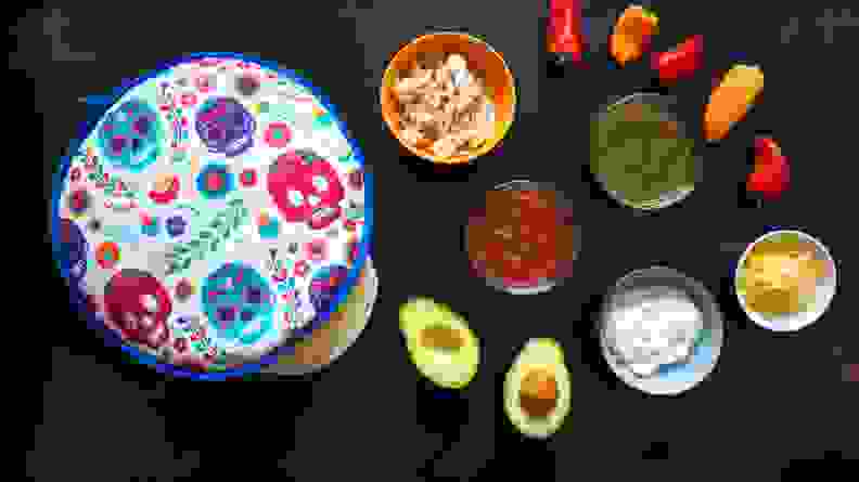 A layout of a tortilla warmer with tortillas peeking out, with taco ingredients like salsa, avocado, and peppers.