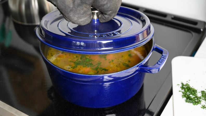 A person lifts the lid off of a blue Dutch oven to reveal chicken soup cooking inside.