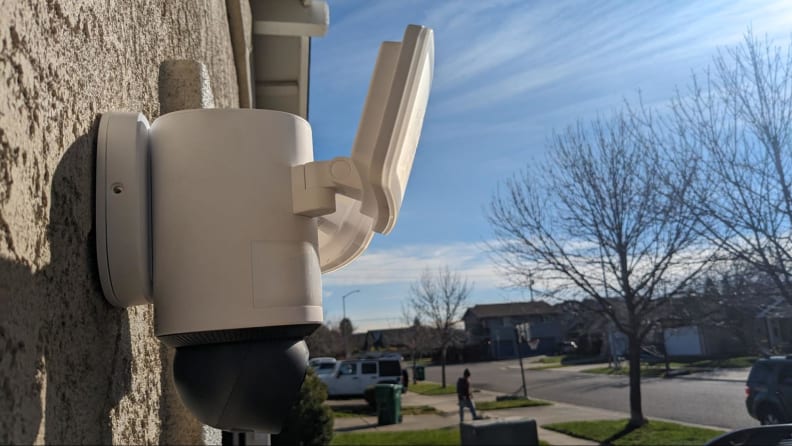 The Eufy Floodlight Camera E340 shown mounted on the exterior of a beige home with a blue sky and a neighborhood in the background.