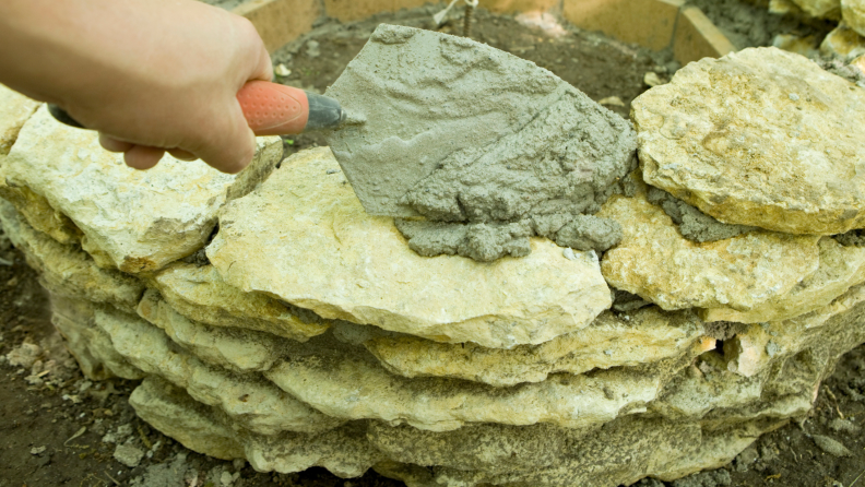 Person using spatula to apply wet cement to concrete stones in order to construct fire pit.