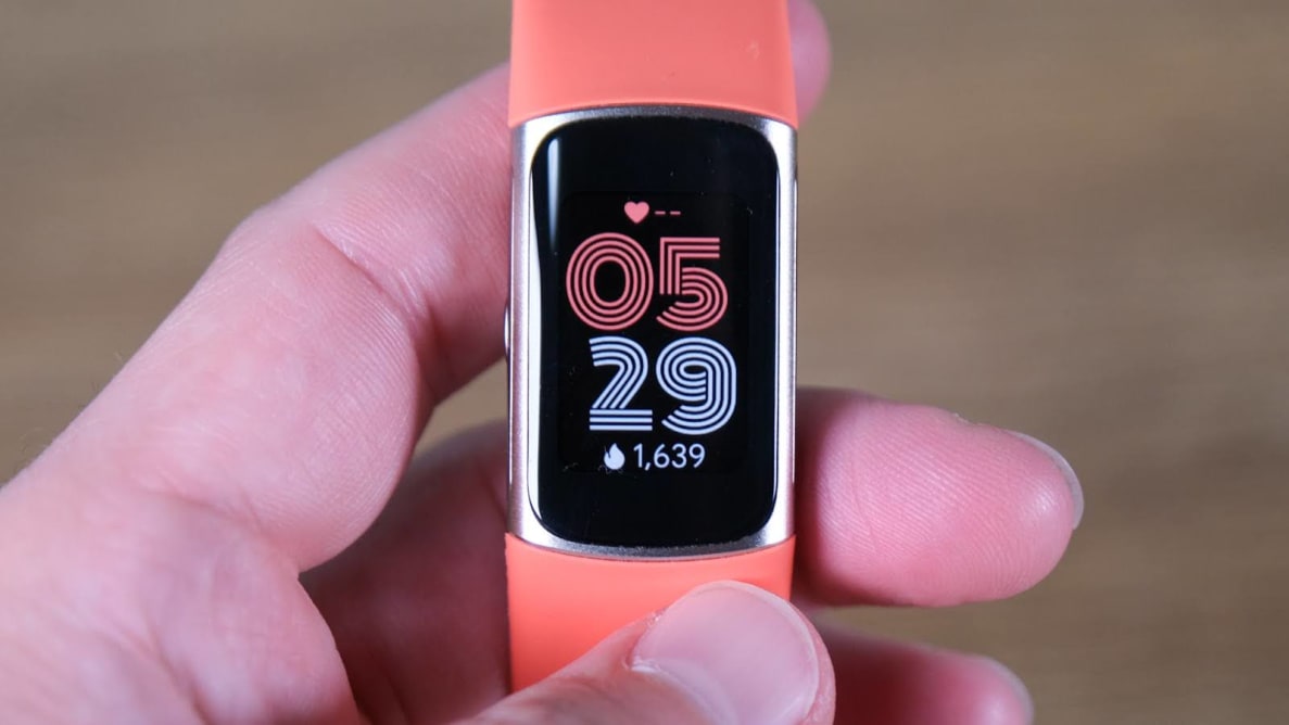 Person holding a Fitbit Charge fitness watch with orange band within their fingers.