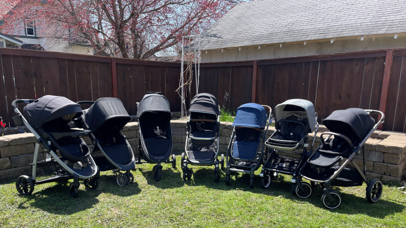 Six different strollers lined up in a backyard.