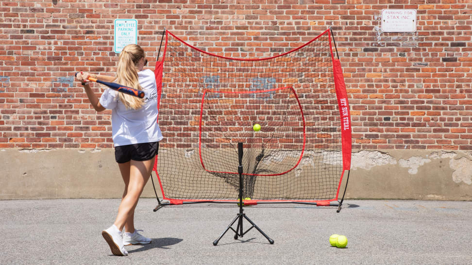 Person in athletic clothing hitting baseball with baseball bat into the Hit Run Steal softball hitting net in front of brick wall outdoors on sunny day.
