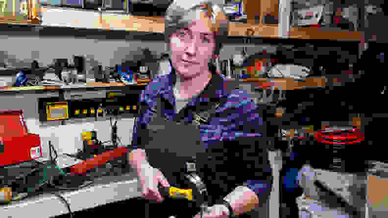 The author stands by her busy workbench, in a work apron, hammer in hand.
