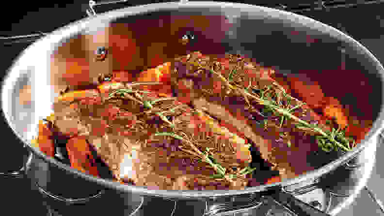A pot on a stove with some meat on top of carrots with rosemary.
