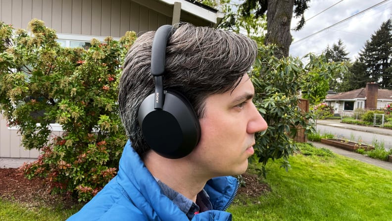 A man in a blue vest listens to the all-black Sony WH-1000XM5 in front of a bush and beige house.