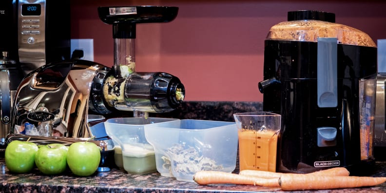 A masticating juicer sits on the left and a centrifugal juicer sits on the right surrounded by produce and cups of juice.