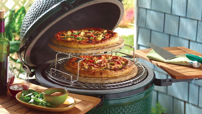 An open Big Green Egg with two pizzas cooking inside, on a tiered grate.