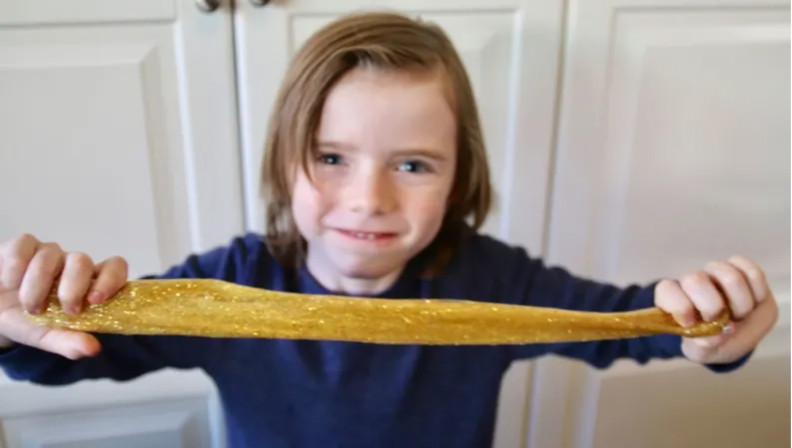 A child in a blue shirt smiles as he stretches yellow slime.