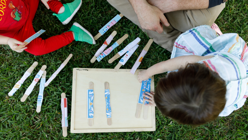 Young boy and girl playing with popsicle sticks outdoors while sitting on green grass.