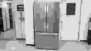 Front view of the silver LG LRFLC2706S Refrigerator against a wall in a white-walled product testing laboratory.