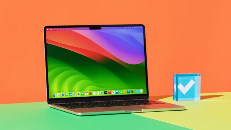 The black laptop sits on a green square on a desk, in front of an orange background