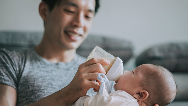 Young father feeding his baby boy son with milk bottle at living room during weekend.