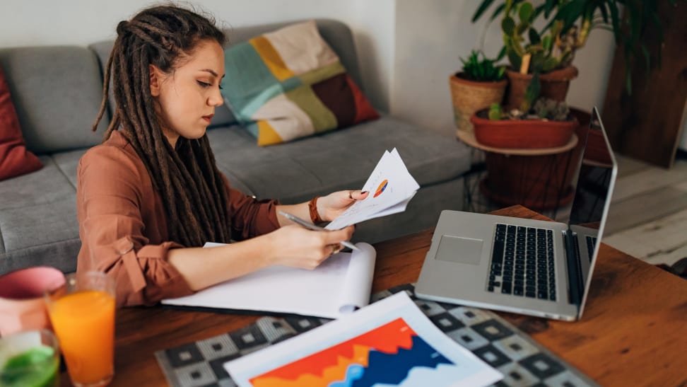 A woman sitting at her coffee table with an open laptop and looking at paperwork