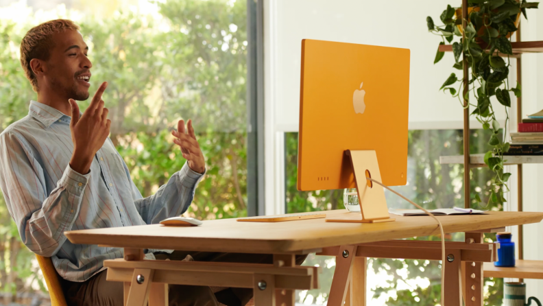 An image of a man sitting at a desk looking at an orange next-gen iMac.