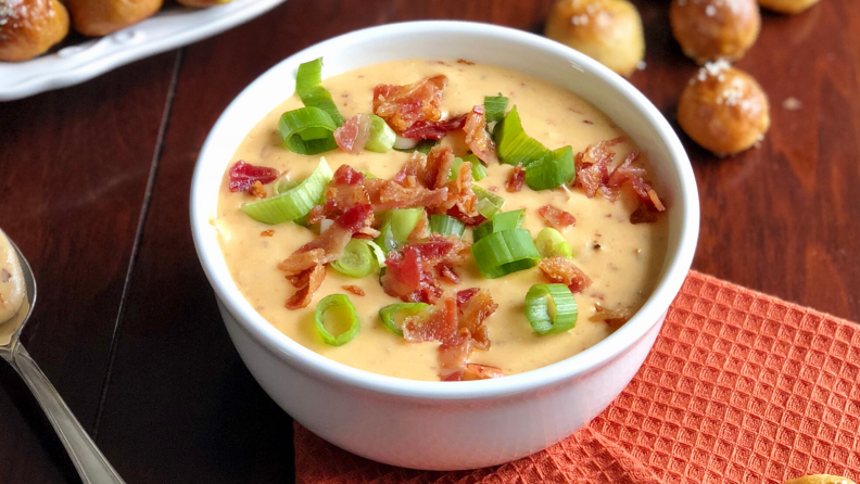 A white bowl filled with a creamy orange dip, topped with bacon bits and fresh scallions. Scattered pretzel bites for dipping in the background.