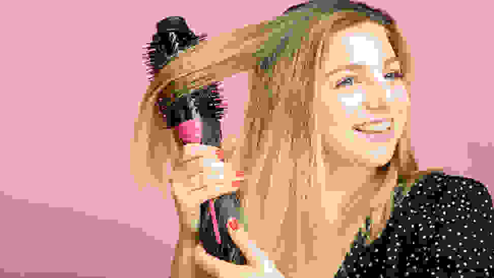 A woman using a hair dryer brush on her blond hair.