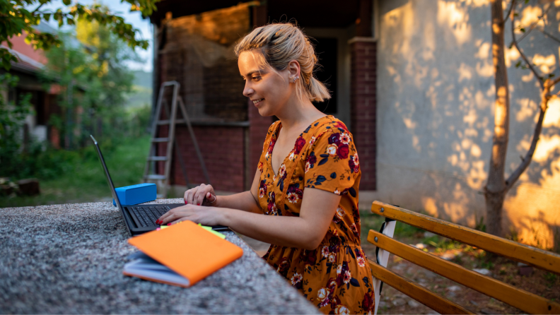 Woman sitting in front of computer outside smiling, next to blue portable speaker and orange notebook.