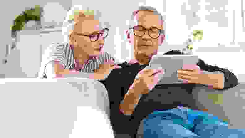 A woman looks over the shoulder of her husband as he sits on an upholstered couch and examines a tablet screen