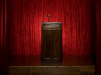 An image of a wooden podium sits on a stage, surrounded by red velvet curtains.