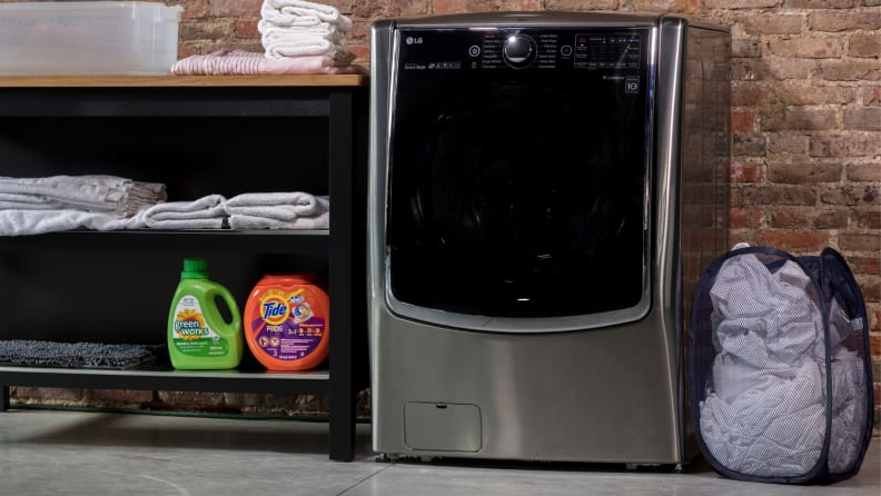 A gray LG WM9000HVA washer sits against a brick wall next to a clothes hamper.