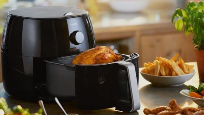 Air fryer on a kitchen counter with a whole chicken inside next to a plate of fries and other food