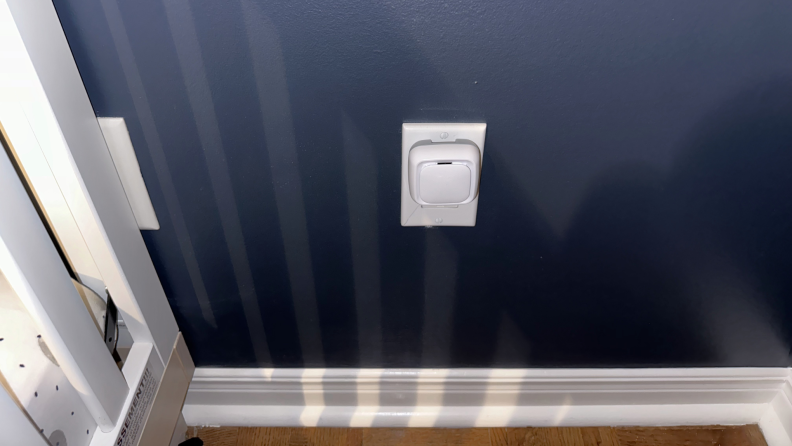Square white smart sensor plugged into wall outlet on navy painted wall.