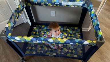 A small toddler sits on top of a patterned mattress inside of the Cosco Funsport Portable Compact Baby Playard.